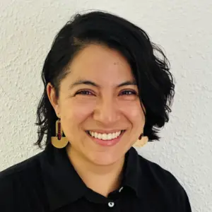 A woman with short, wavy black hair smiles broadly. She wears large gold geometric earrings and a black collared shirt. Textured white wall in the background.